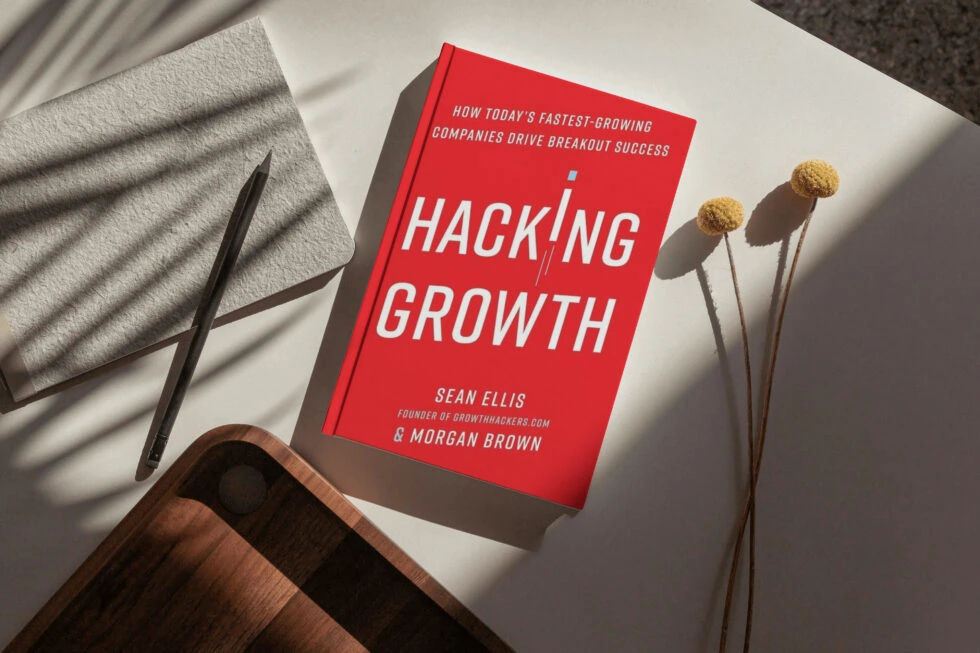 A photo showing the book "hacking growth" by sean ellis and morgan brown, along with a pen, a cozy blanket, and dried flowers, creating a cozy workspace setting.