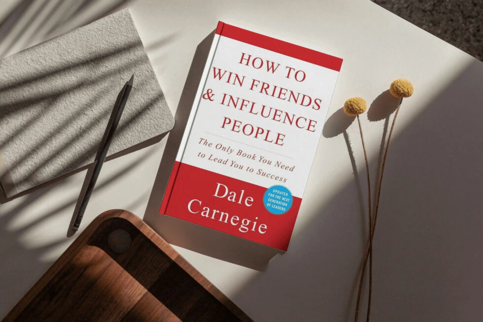 A classic book titled "how to win friends & influence people" by dale carnegie, accompanied by a pen, dried flowers, and a wooden surface.