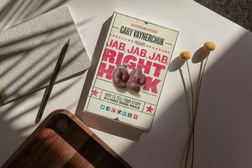 A book titled "jab, jab, jab, right hook" by gary vaynerchuk, along with boxing gloves and dried flower stems, lying on a white surface with a pen and a towel.