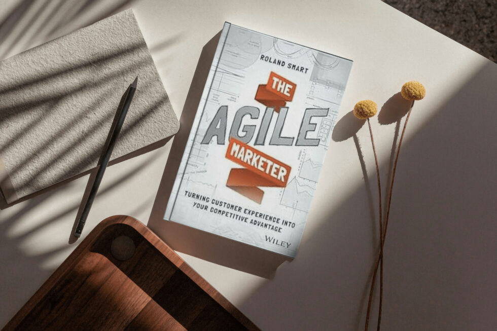 A book titled "the agile marketer" by roland smart, lying on a surface alongside a pen, towel, and dried flower stems.