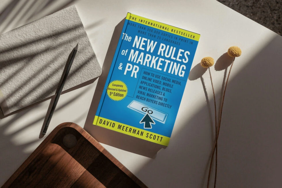 A book titled "the new rules of marketing & pr" by david meerman scott, along with a pen, dried flowers, and a wooden surface, highlighting social media and digital marketing strategies.