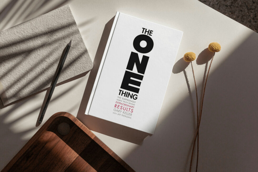 A book titled "the one thing" by gary keller and jay papasan, along with a pen, dried flowers, and a wooden surface, focusing on achieving extraordinary results through a simplified approach.
