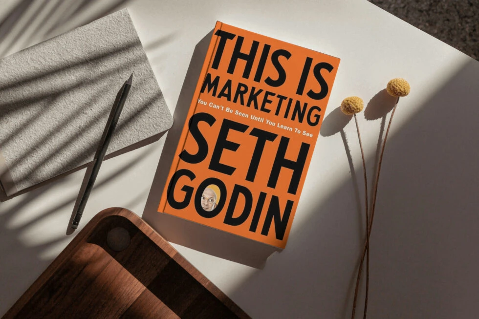 The book "this is marketing" by seth godin lying on a surface alongside a pen, towel, and dried flower stems.