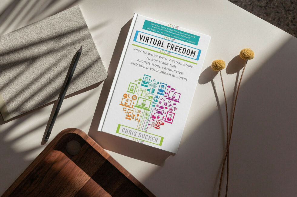 A book titled "virtual freedom" by chris ducker, along with a pen, towel, and dried flower stems arranged on a white surface.