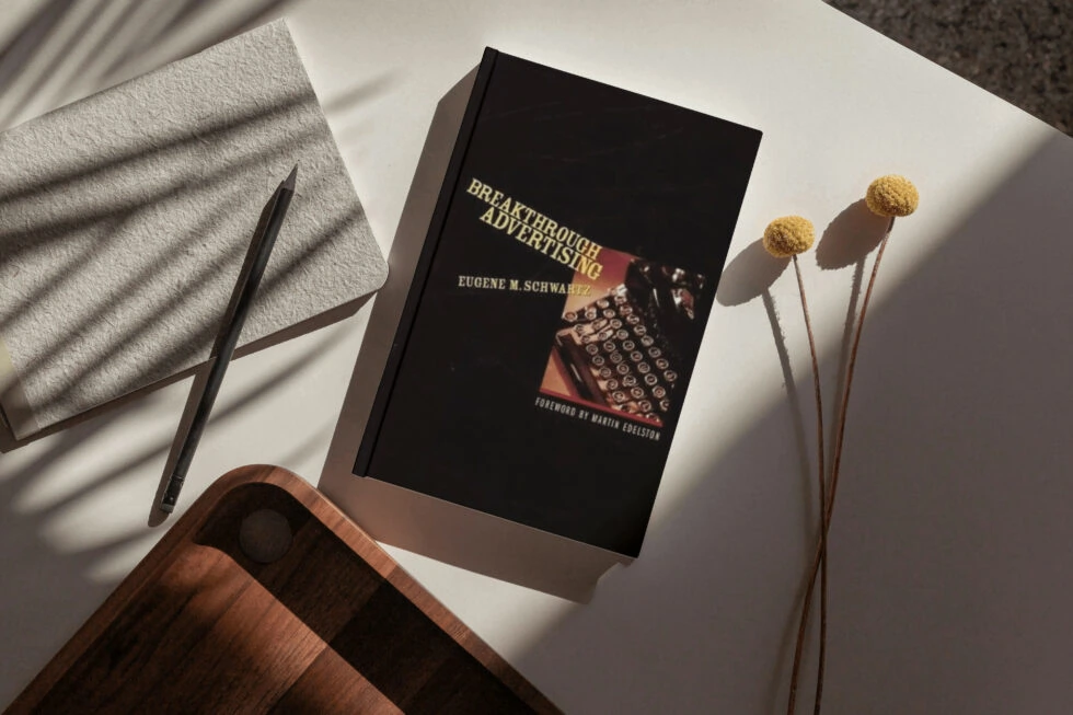 A black hardcover book titled "breakthrough advertising" by eugene m. Schwartz, a renowned guide on effective advertising practices, surrounded by a pen, dried flowers, and a cozy blanket, creating a thoughtful reading environment. Image caption: eugene schwartz's "breakthrough advertising" is consider