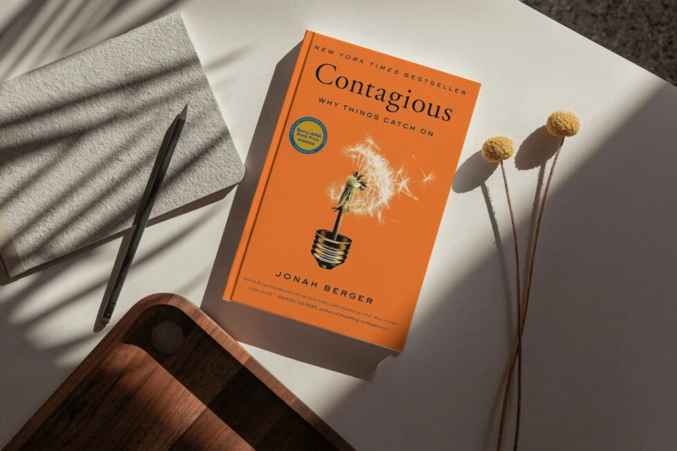 A copy of the new york times bestseller "contagious: why things catch on" by jonah berger, along with a pen, dried flowers, and a cozy blanket creating a reading nook.