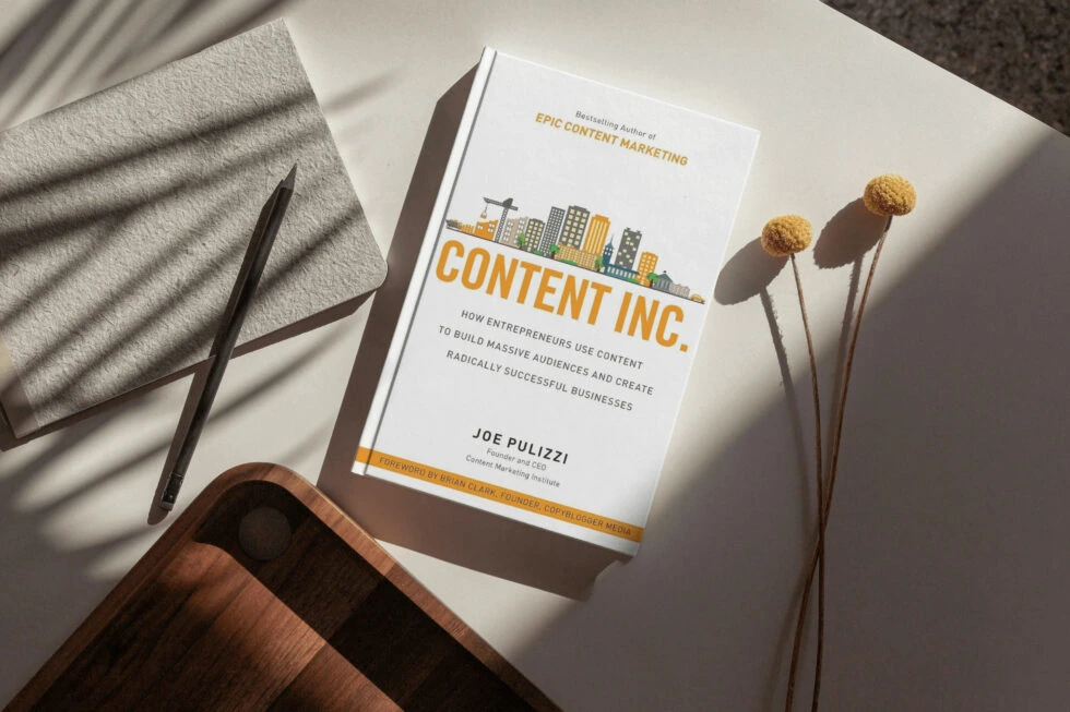 A copy of the book "content inc. " by joe pulizzi, which discusses how entrepreneurs can use content to build massive audiences and create successful businesses, along with a pen and decorative dried flowers on a textured surface.