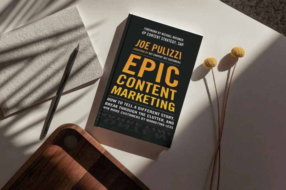 A book titled "epic content marketing" by joe pulizzi, with a foreword by michael brenner of sap, lying on a table with a pen and dried flower stems, focusing on storytelling to engage customers