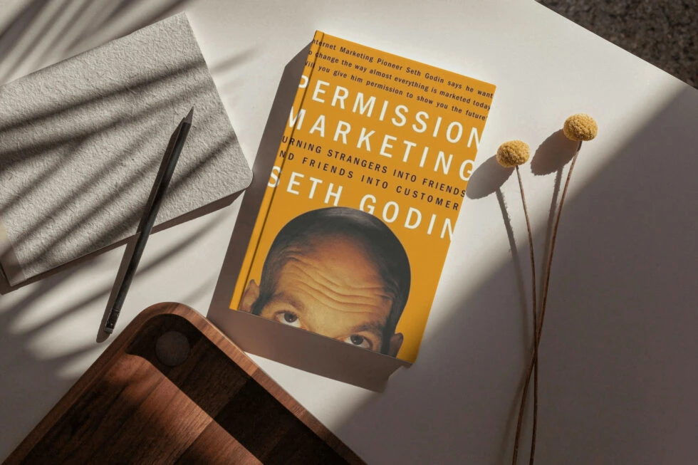 A marketing book titled "permission marketing" by seth godin, an internet marketing pioneer, lying on a table with a pen and dried flower stems